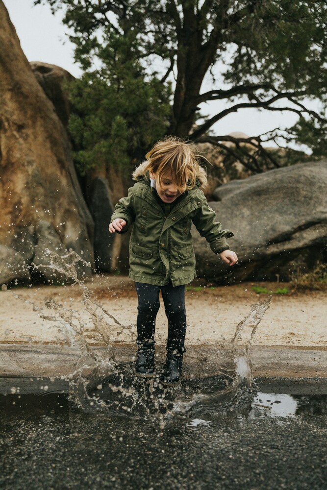 Ein kleines Kind in einem grünen Parka springt in eine Pfütze und spritzt dabei Wasser hoch.