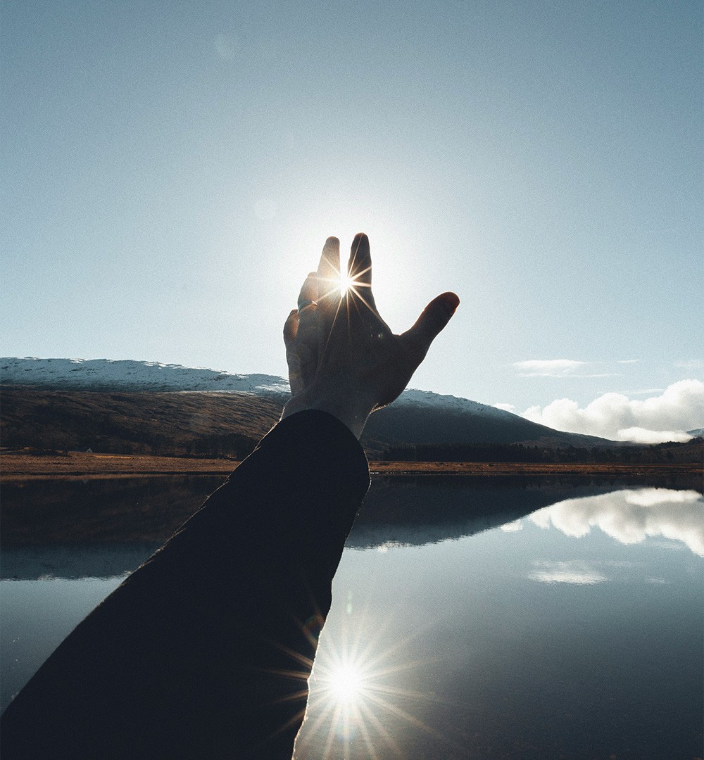 Ein Arm mit einer Hand, die sich gegen die Sonne erhebt, vor einer ruhigen Wasserfläche mit Spiegelung und einem Berg im Hintergrund.